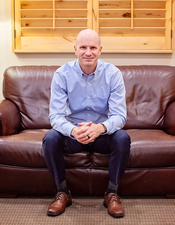 A dentist in a light blue button-down shirt seated on a brown leather couch.